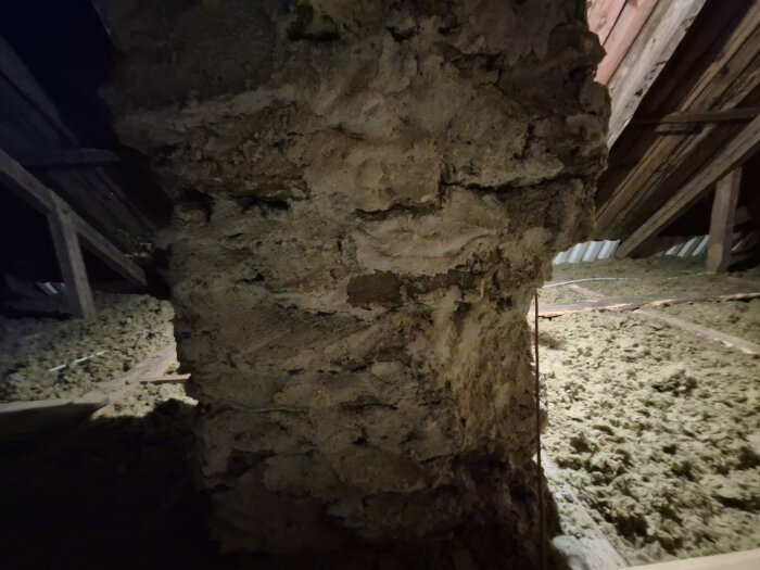 Ett loft med en murad pelare i mitten, träbalkar och isoleringsmaterial mellan balkarna. Diskuteras att lägga golv för förvaring.