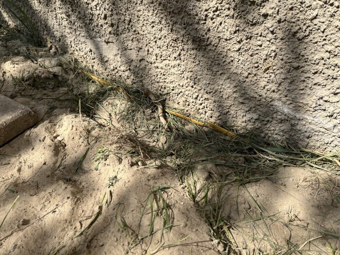 Gul dräneringsplast syns i marken vid husets grund, omgiven av sand och grässtrån, och ligger delvis begravd i jorden.
