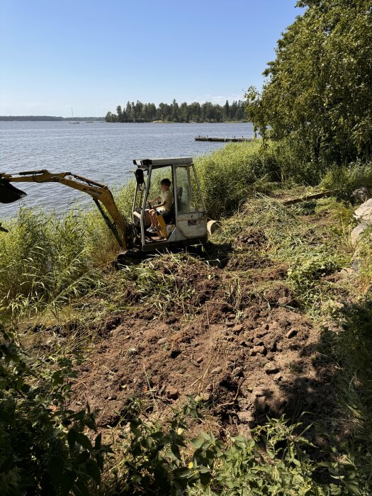 En person kör en liten grävare vid en sjökant och har grävt upp marken runtomkring. Gräset är nedtrampat och marken synlig. I bakgrunden syns sjön och en brygga.