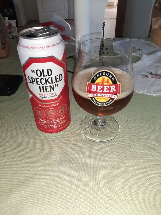 En öppnad burk Old Speckled Hen English Pale Ale och ett glas med öl, placerade på ett bord med en ljus duk.