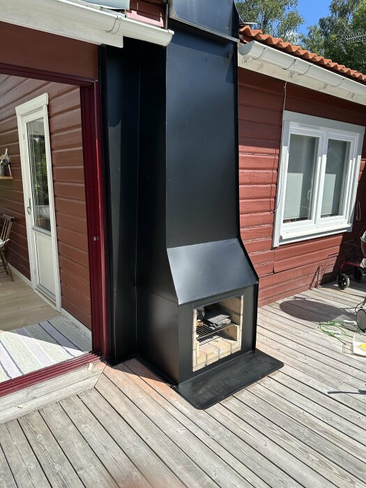 Utmomhusbild på en nyinstallerad kamin inklädd med svart plåt, intill en röd träfasad med vitmålande fönsterkarmar och tegelpannor på taket.