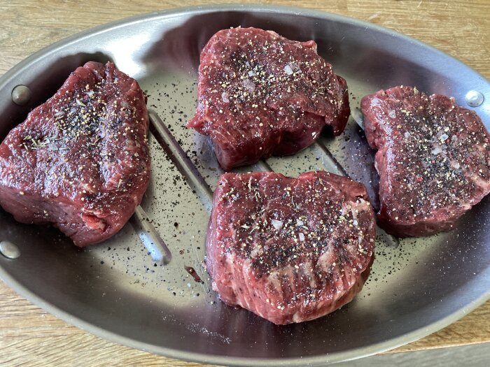 Fyra bitar okokt kött kryddade med salt och peppar ligger på en metallbricka, redo för grillning.
