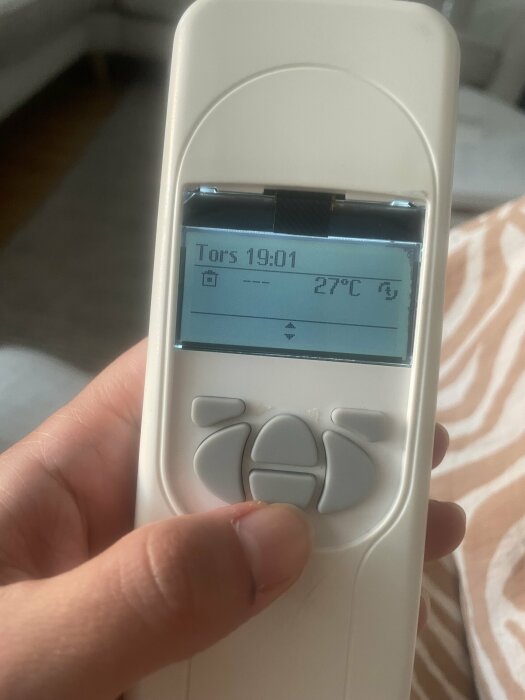 Hand som håller en fjärrkontroll med display som visar "Tors 19:01" och "27 °C". Kontrollens knappar och bakgrund delvis synliga.