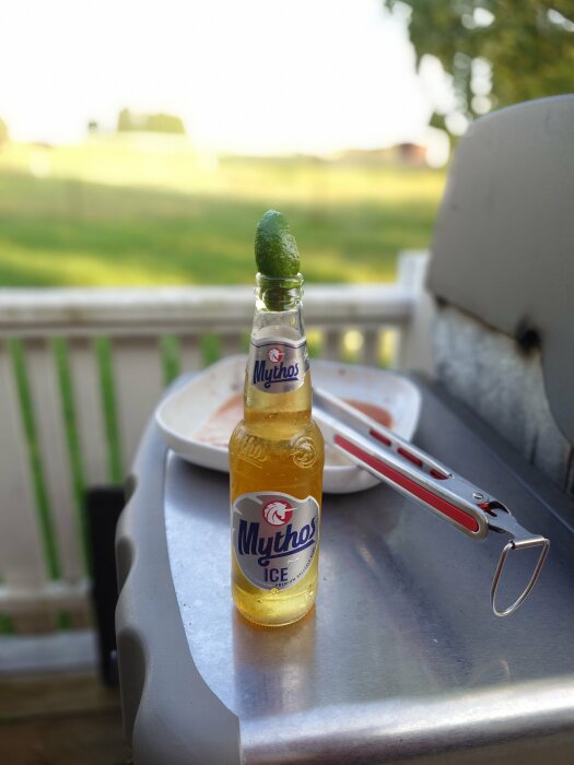 En flaska Mythos Ice öl med en limekil på toppen står på ett grillbord bredvid en grilltång och en tom tallrik, med en utsikt över en grön gräsmatta i bakgrunden.