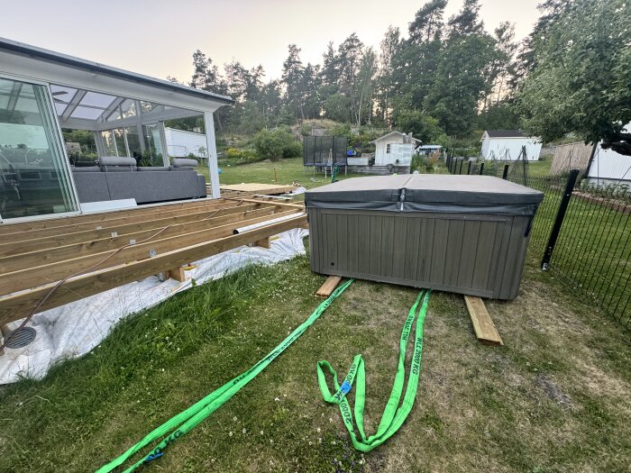 Ett stort spabad står på träplankor bredvid ett hus med terrass under byggnation. Gröna lyftslingor ligger på gräsmattan bredvid spabadet.