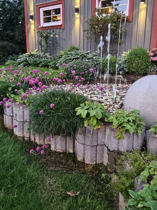 Trädgård med blommande rabatter, en fontän och kantstenar med jordgubbs- och smultronplantor framför ett hus.