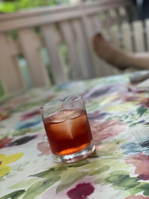Ett glas Negroni med is på ett färgglatt blommigt bord, en fot syns i bakgrunden.