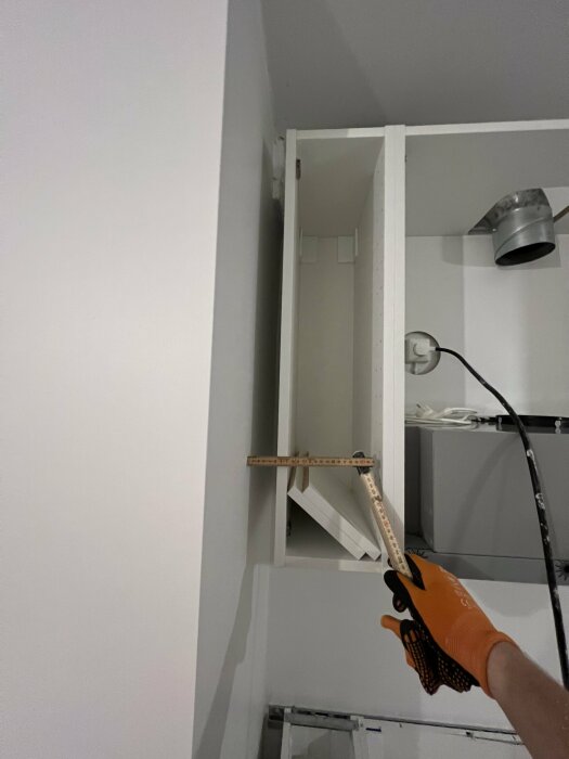 En hand med orange handske använder ett mätverktyg för att justera ett väggskåp i ett kök under renovering. Rör och kabel syns i bakgrunden.