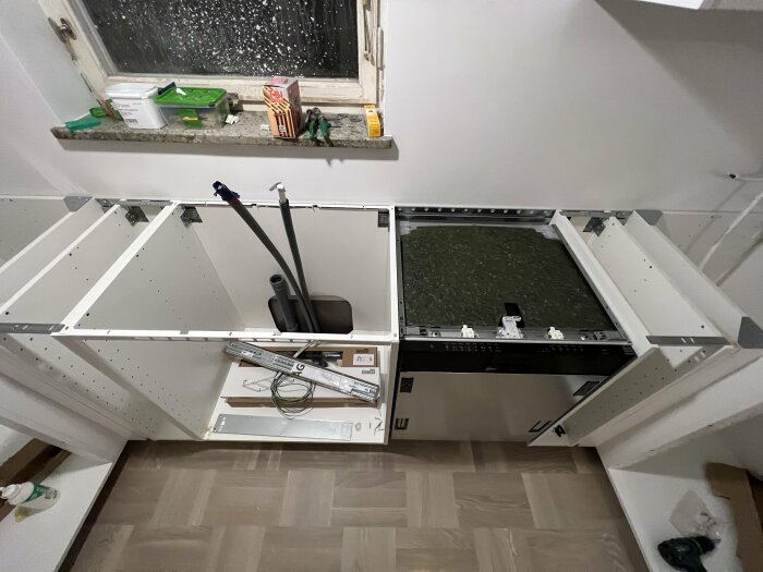 En halvt färdigställd köksrenovering med vita skåpstommar, handverktyg på fönsterbrädan och rörledningar som förbereds för montering av bänkskiva.