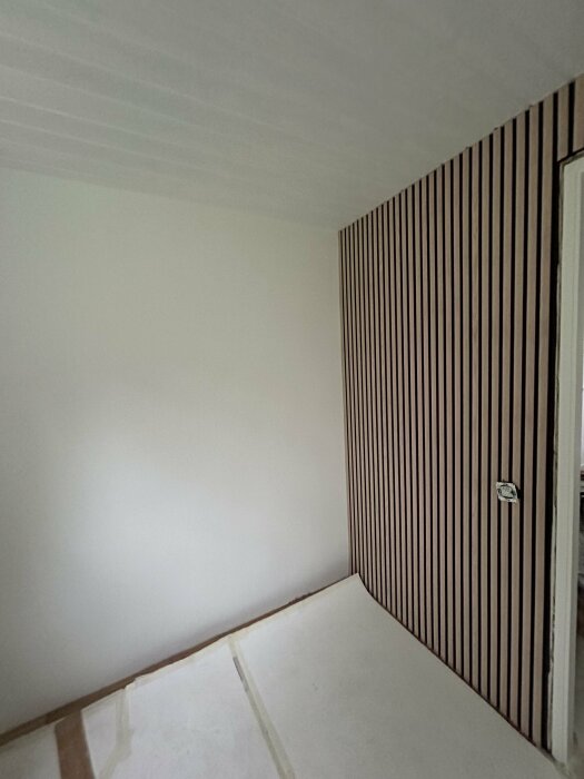 Ljusgrått rum med en vit vägg och en vägg med vertikala träpaneler. Golvet är täckt av skyddspapper och en vit filt syns på golvet.