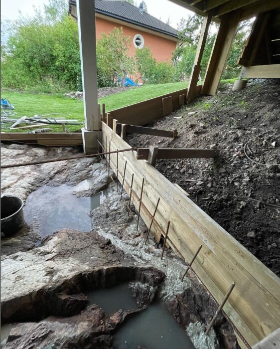 Pågående arbete med byggandet av en stödmur under en altan, som visar armeringsjärn och en form för betonggjutning på stenig mark och intilliggande gräsmatta.