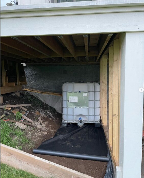 IBC-tank placerad under en altan, omgiven av byggmaterial och stödmurar. Markavloppsrör löper längs marken bredvid tanken.
