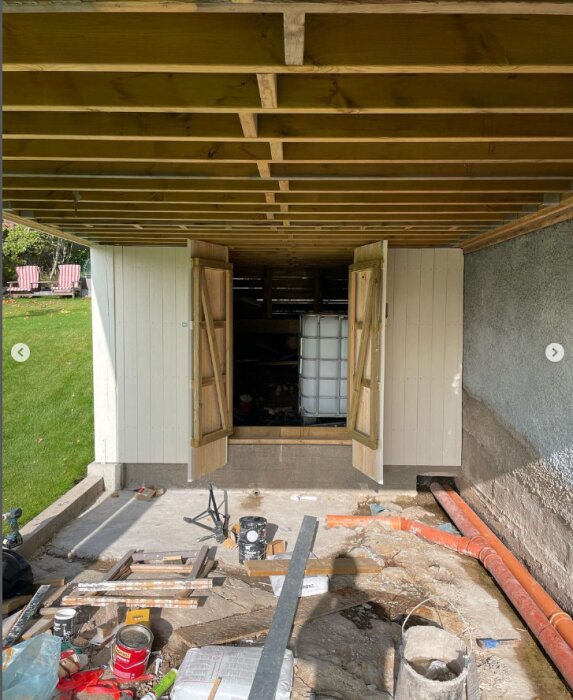 Under en nybyggd altan ses en upphöjd plattform med en stödvägg, panelklädda väggar och två öppna dörrar som leder in till förvaring med en IBC-tank. Byggmaterial och verktyg ligger utspridda.