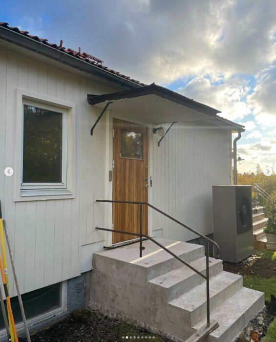 Renoverad ytterdörr av trä i en vitmålad husfasad, med trappa av betong block och enkel metallräcke framför dörren.