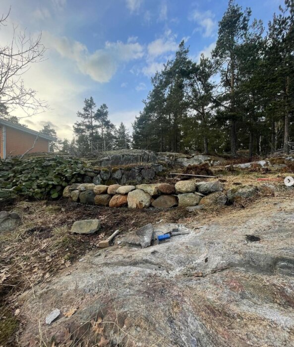 Låg stenmur byggd med stora stenar på en tomt med träd i bakgrunden, verktyg och byggmaterial ligger på marken framför muren.