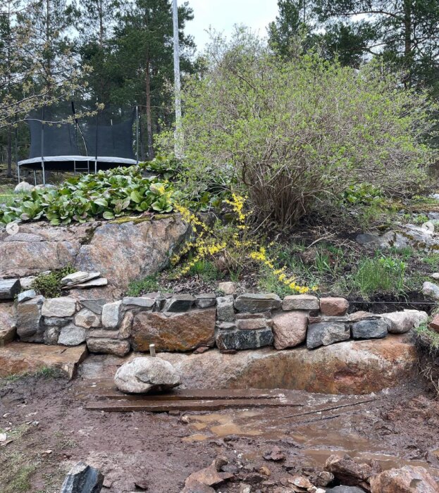 Byggd stenmursgrund med stenar i olika storlekar och färger, omgiven av grönska och växter, delvis färdig konstruktion.