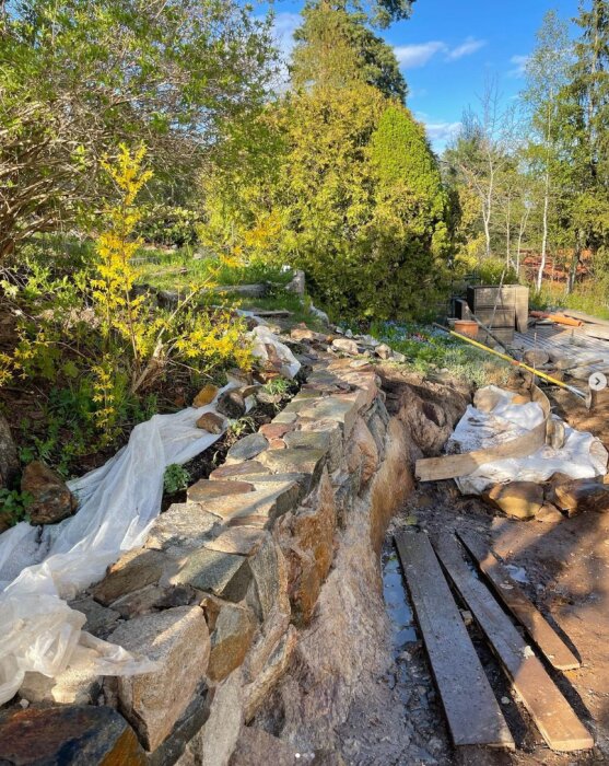 En halffärdig stenmur som löper längs en sluttning med plastöverdrag, bräder och byggmaterial runtom. Vegetation och träd i bakgrunden.