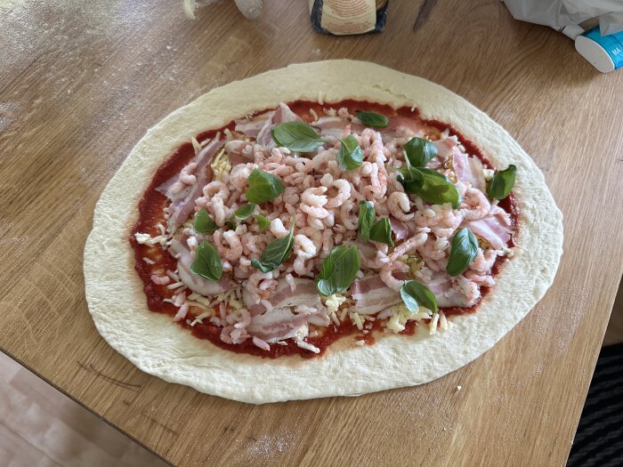 En hemmagjord pizza på trämaterial med tomatsås, riven ost, räkor, bacon och färska basilikablad som pålägg.