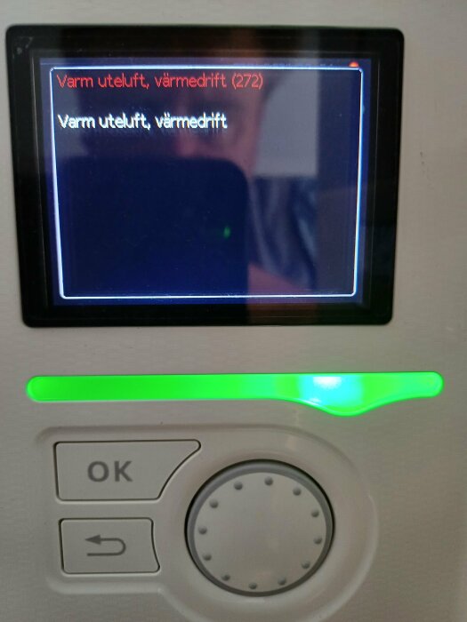 Display på en värmepumpskontrollpanel som visar meddelandet "Varm uteluft, värmedrift (272)" och knapp med texten "OK" samt ett reglage med grön bakgrundsbelysning.