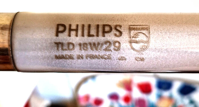 Närbild på ett äldre Philips-lysrör med texten "PHILIPS TLD 18W/29 Made in France" synligt på ytan. Färgglad bakgrund delvis synlig i nedre delen av bilden.