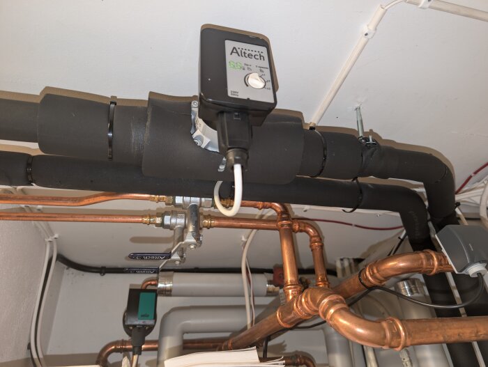 Vattenrör med förgreningar och ventiler i en källare. Ett rör är isolerat och en Frico-enhet och en Wilo-pump är installerade på vattenledningar.
