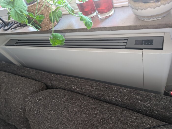 Vit Frico fläktkonvektor installerad bakom en soffa, under en fönsterbräda med flera växter och glasburkar.