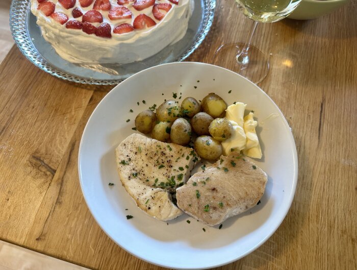 Tallrik med tonfisk, färskpotatis och smör vid sidan; jordgubbstårta i bakgrunden och glas med vit dryck bredvid.