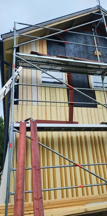 Byggställning vid husvägg med blandad panel, omålad, grundmålad och slutfärgad, under arbete; två rödmålade plankor står lutade mot ställningen.