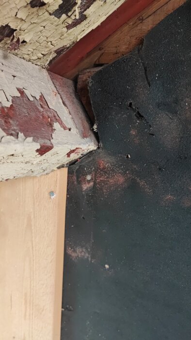 Närbild på hörn av husvägg med målad panel, svarta plattor, och skadad färg på träet.