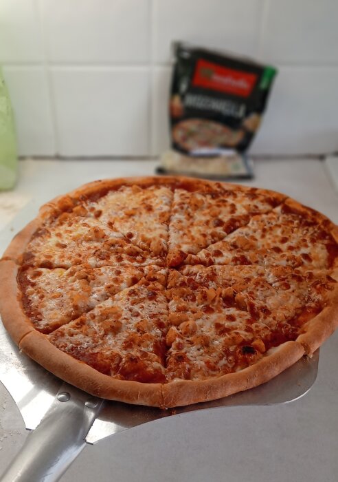 En hemlagad ostpizza på en pizzaspade, bakgrund med kök och påse riven mozzarellaost.