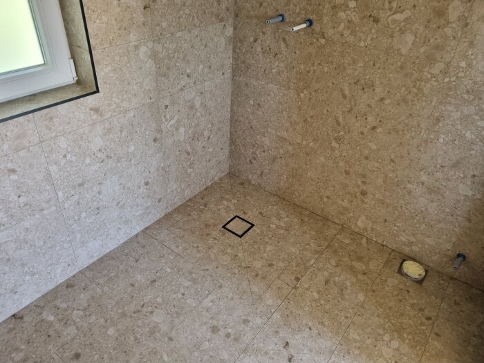 Kakelbeklätt badrum med fönster och golvbrunn, 30x60 cm plattor, vattenrör och placering för toalett eller duschmunstycke.