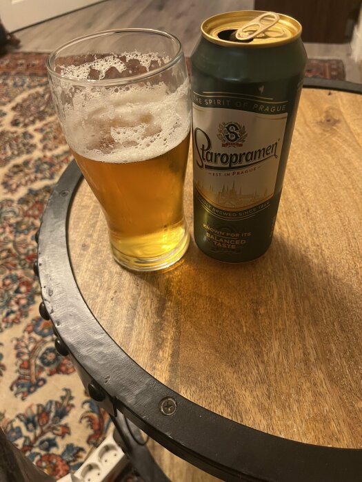 En öppnad burk Staropramen-öl och ett halvt uppdrucket ölglas står på ett runt träbord med metallkant. Ett mönstrat matt hörn syns i bakgrunden.