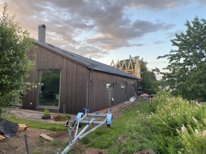 Nybyggt hus med träfasad, takstolar har kommit upp på ena änden, omgivande grönska, en blå trailerkonstruktion syns i förgrunden under en mulen himmel.
