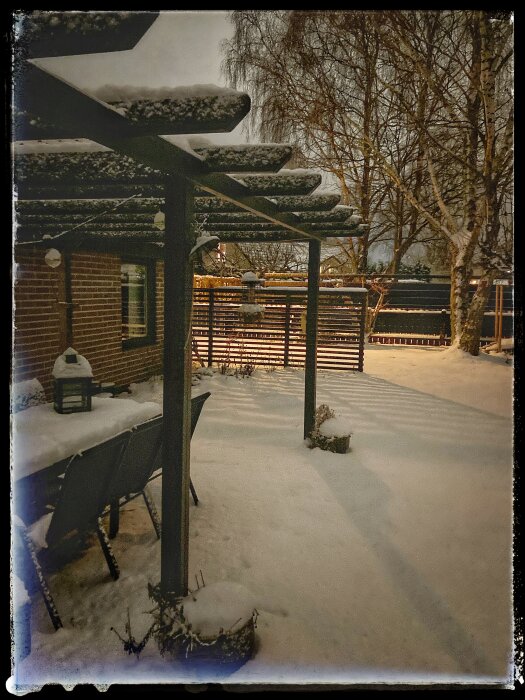 Pergola i en trädgård under vintern, täckt med snö, omgiven av snötäckta träd och utemöbler.