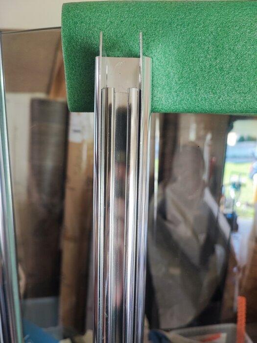 Närbild på övre delen av en rundad duschdörr i glas monterad i en metallram, med skyddande grönt material ovanpå. Bakgrund med suddiga föremål och en spegling.