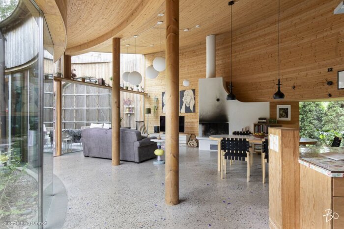 Modernt vardagsrum med träpaneler, stor fönstervägg, grå soffa, öppen spis och matbord, samt öppen planlösning mot köket med trädetaljer och pendantlampor.