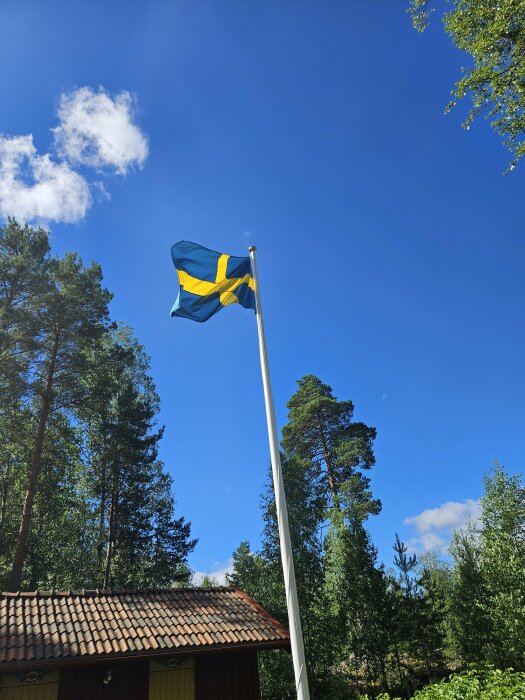 Svenska flaggan vajar på en flaggstång framför ett rött hus med tegeltak, omgivet av grönskande träd under en klarblå himmel med några vita moln.