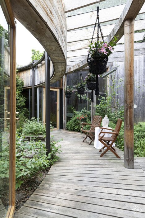 Grönskande trädgårdsgång med träöverdrag, stolar och bord, samt hängande blommor under ett delvis täckt tak.
