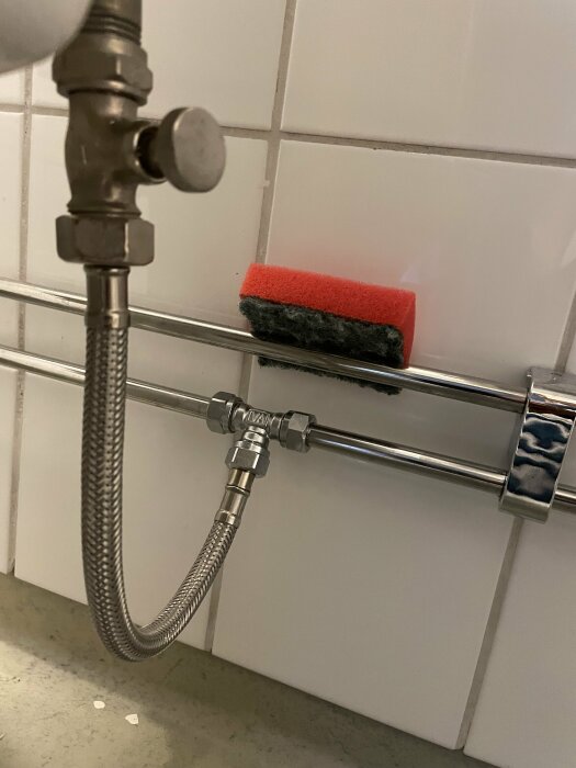 Ventil för vattentillförsel till toalett monterad på vägg, med en flexibelt metallslang ansluten. Röd svamp fastsatt på rör.
