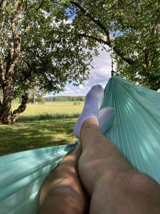 Person som kopplar av i en hängmatta på en solig dag, med utsikt över en grön trädgård och blå himmel genom träden. Delen av benen synliga i bild.