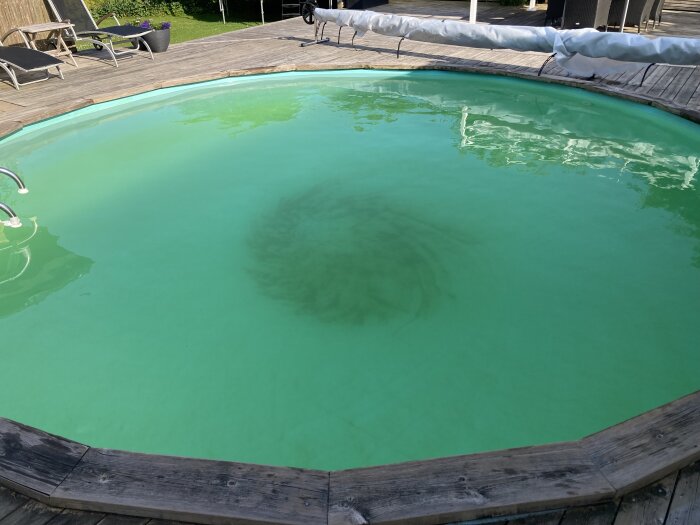 Rund pool med grumligt och grönt vatten. Det syns en mörkare ring i mitten av poolens botten. I bakgrunden finns en träterrass och solstolar.