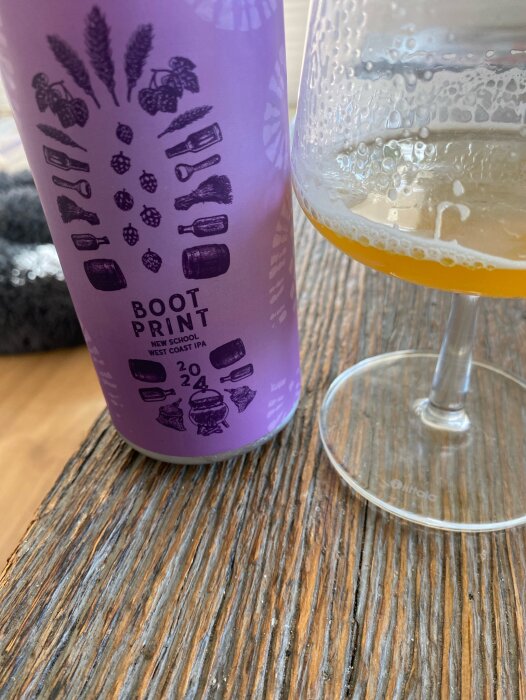 Lila burk med etiketten "Boot Print New School West Coast IPA" bredvid ett fyllt glas på ett bord.