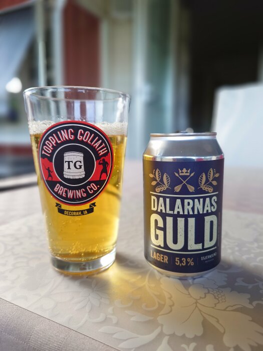 Ett glas öl märkt "Toppling Goliath Brewing Co." bredvid en burk öl med namnet "Dalarnas Guld" på ett bord med dekorativ duk.