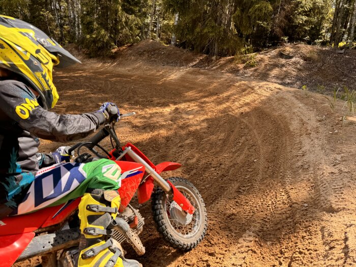 Person på motocross cykel med färgglada kläder och hjälm på en lerig bana i skogen.