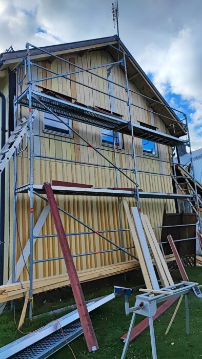 Husrenovering med byggnadsställningar och träpaneler monteras på fasaden. Verktyg och virke uppställt på gräsmattan under ställningen.