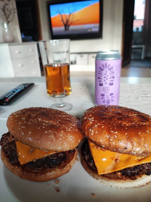 Två hamburgare med smält ost på en tallrik, ett glas med dryck, en lila läskburk och en TV i bakgrunden som visar en ökenbild.