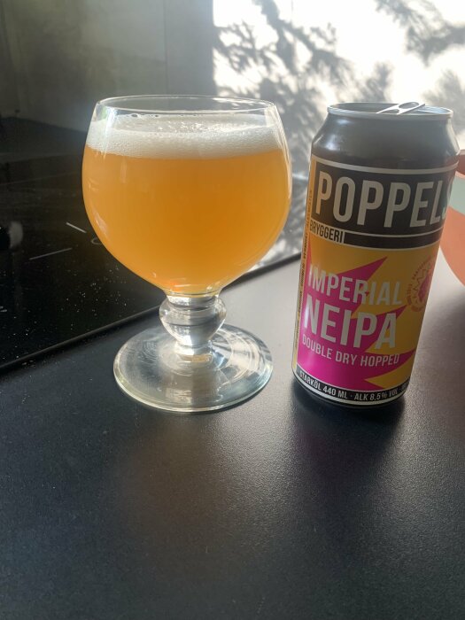 Ett glas fylld med ljus öl och en ölburk från Poppels Bryggeri märkt med "Imperial NEIPA Double Dry Hopped" placerad på ett svart bord.