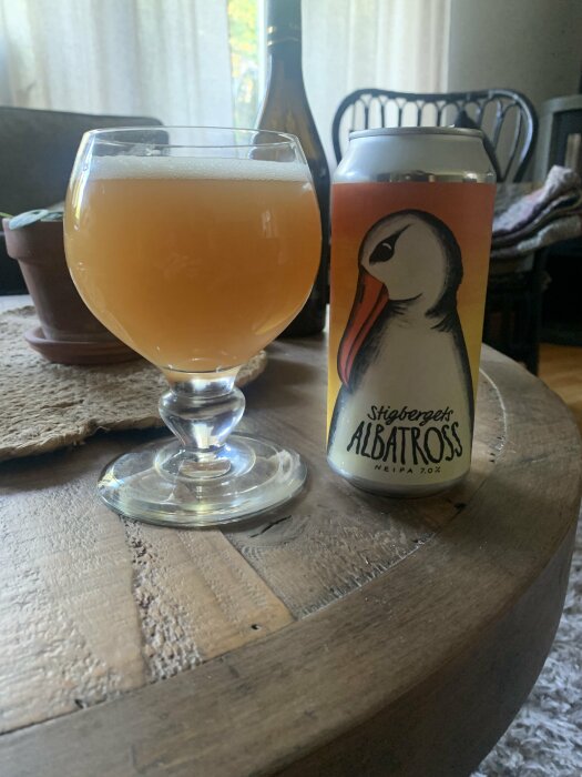 Ett glas Stigbergets Albatross NEIPA och en ölburk som står på ett rustikt träbord, med en krukväxt och en vinflaska i bakgrunden.