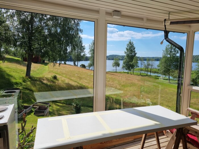Provisorisk bänkskiva i träverkstad med utsikt över grönska och sjö genom stora fönster i ett uterum.