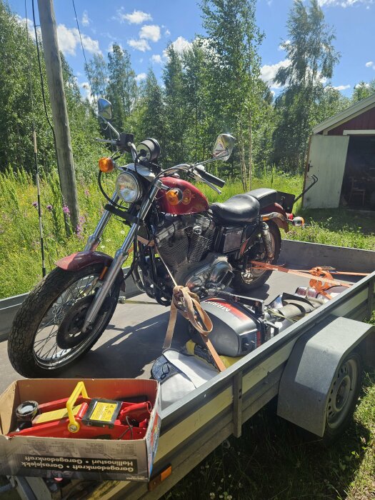 Motorcykel fastspänd på ett släp med verktygslåda och generator omgiven av grönska och ett rött uthus i bakgrunden.